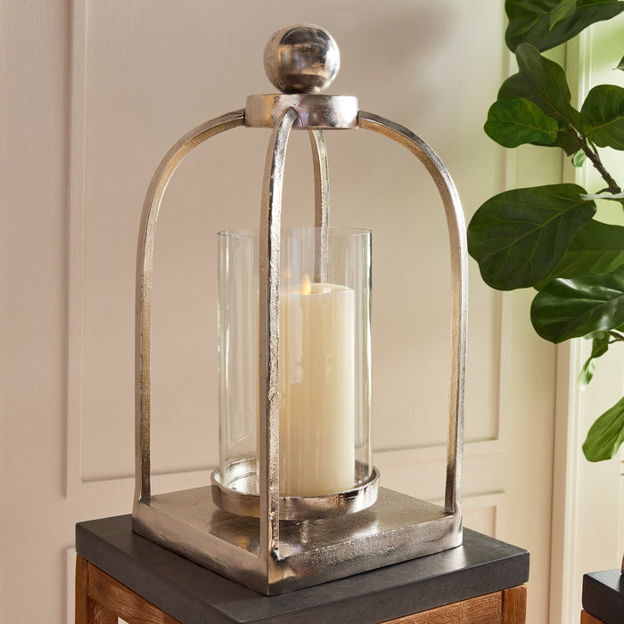 Design Shop Hollis lantern with metallic silver frame, glass cylindrical enclosure, large ivory candle, on dark wood base.