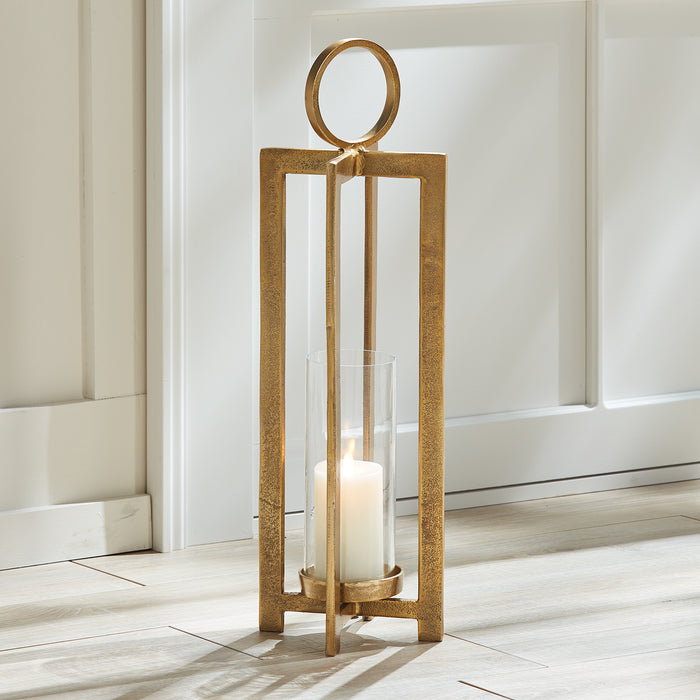Gold-tone metal lantern with circular top ring, glass cylinder, and white candle inside on light wooden floor by