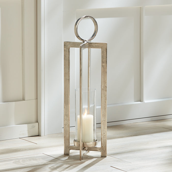Tall rectangular metal lantern with a circular handle, silver finish, glass cylinder candle holder, on light wood floor near