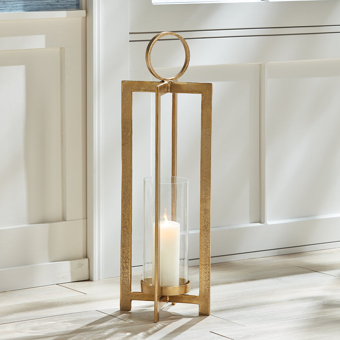 Gold lantern with rectangular metal frame and circular top ring, clear glass cylinder around a white pillar candle, standing
