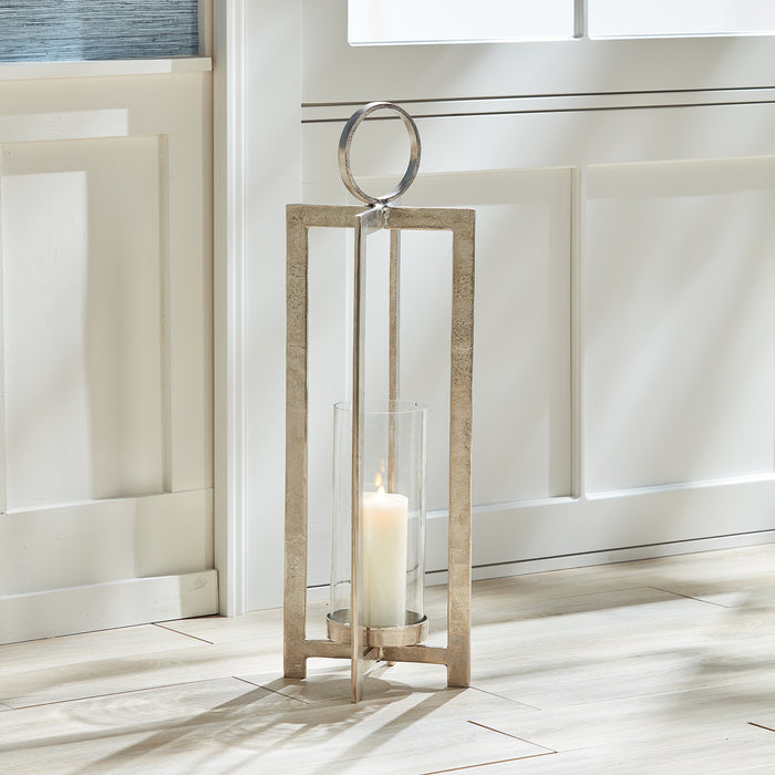 Large lantern with metallic silver frame, circular ring top, glass panels, and a lit pillar candle inside on a pale wooden