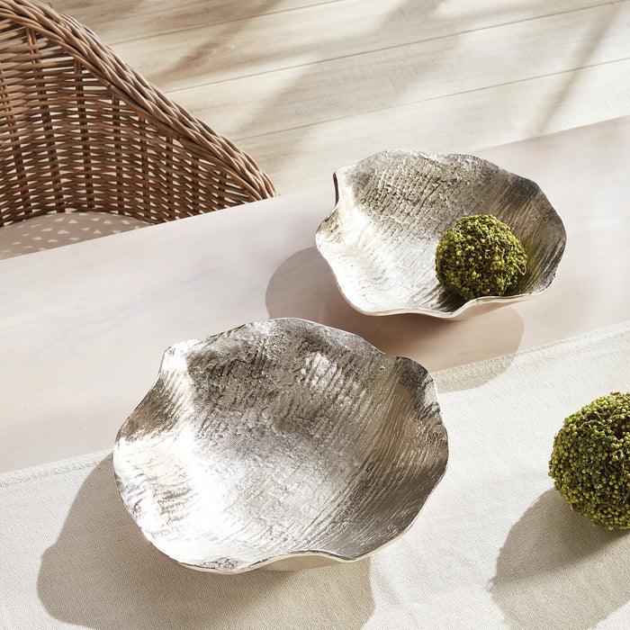 Two textured silver decorative bowls with organic wavy edges on a light table, each containing a green moss ball.