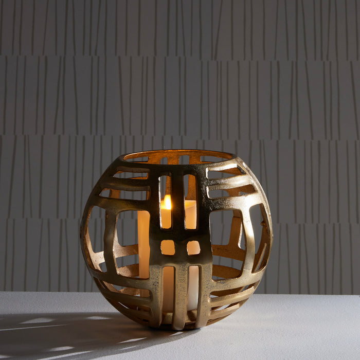 Bronze spherical lattice candle holder with warm flame glow, open geometric cutouts, against a textured gray wall.