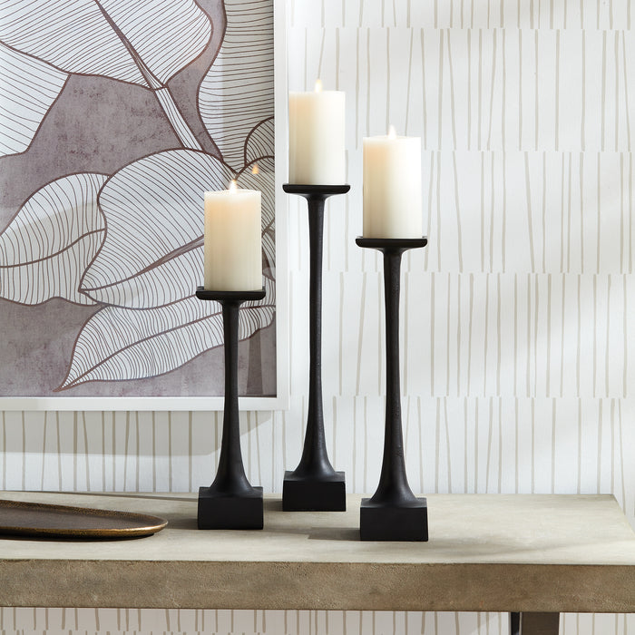 Three black metal candlesticks of varying heights with cream pillar candles on a beige surface against patterned wallpaper.
