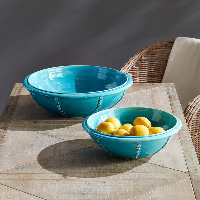 Two turquoise ceramic bowls with stitched side handles on a wooden table; one filled with lemons, one empty, wicker chair in