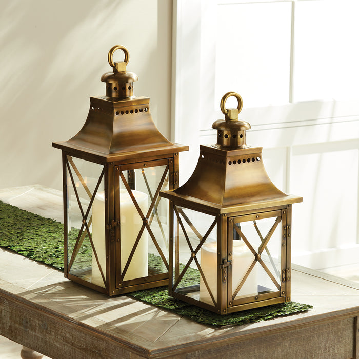 Two bronze lanterns with glass panes and white candles inside, decorative crossbars, atop a wooden table with a green runner.