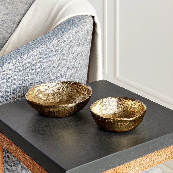 Two gold-toned hammered bowls on a dark gray tabletop, with a blue-gray chair and light wall in background.
