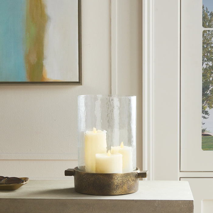 Brass-toned cylindrical candle holder with a hammered texture, glass hurricane, three ivory pillar candles, on a light