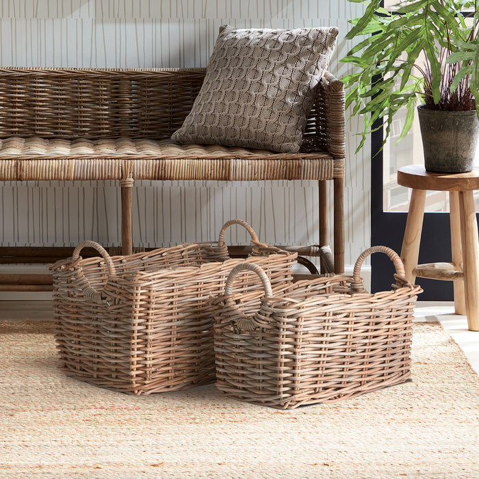 Two rectangular woven rattan baskets with handles on a natural jute rug, in front of wicker sofa and plant.