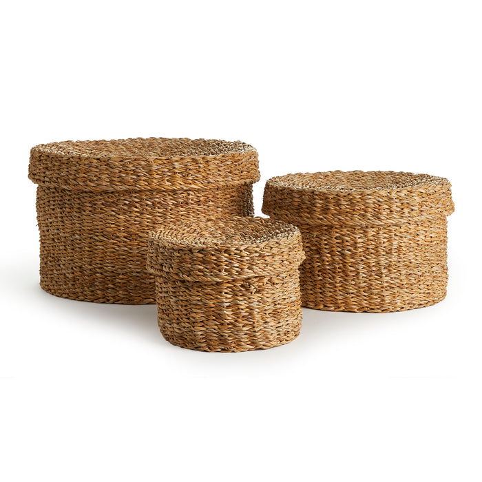 Three round seagrass baskets with lids in natural brown hues, varying sizes, woven texture, plain white background.