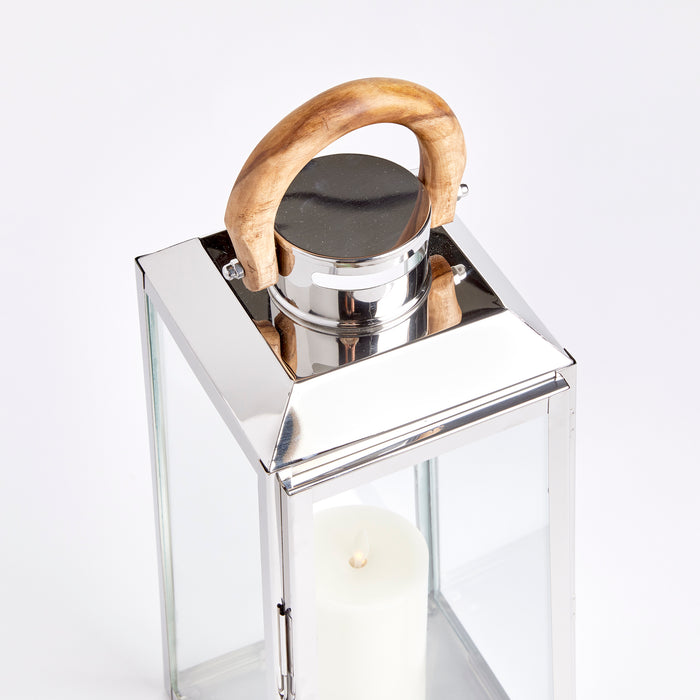 Large chrome and glass outdoor lantern with a curved wooden handle and a lit white pillar candle inside, against a light