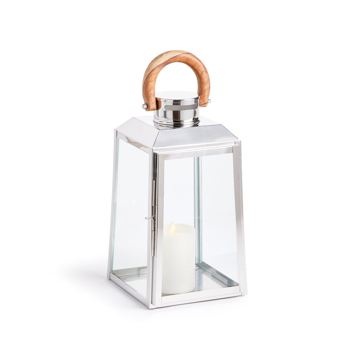 Silver metal lantern with glass sides, wooden curved handle, and lit white pillar candle inside against white background.