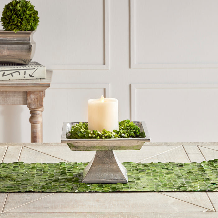 A square, brushed nickel decorative pedestal tray with a candle and greenery on top, placed on a green textured surface.