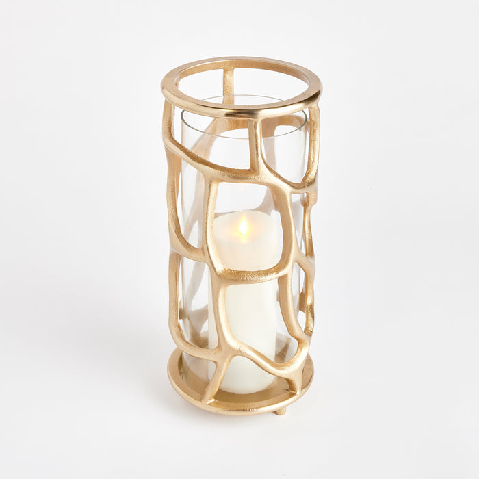 Gold-tone geometric cage lantern with glass side panels surrounding a white pillar candle inside, on a white background.
