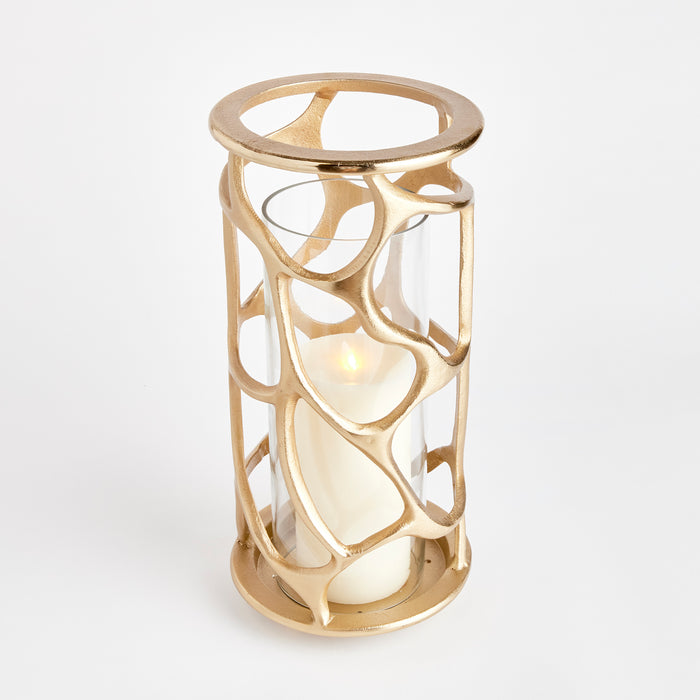 Gold-toned cylindrical hurricane lantern with an abstract open-metal lattice cage around a white pillar candle, glass core