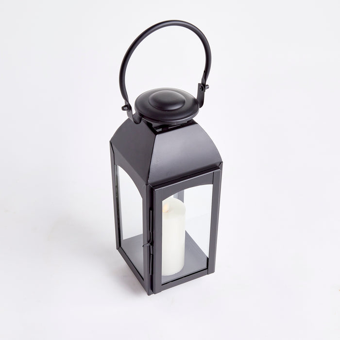 Black metal outdoor lantern with glass panels and a white pillar candle inside, ring handle on top, against a white