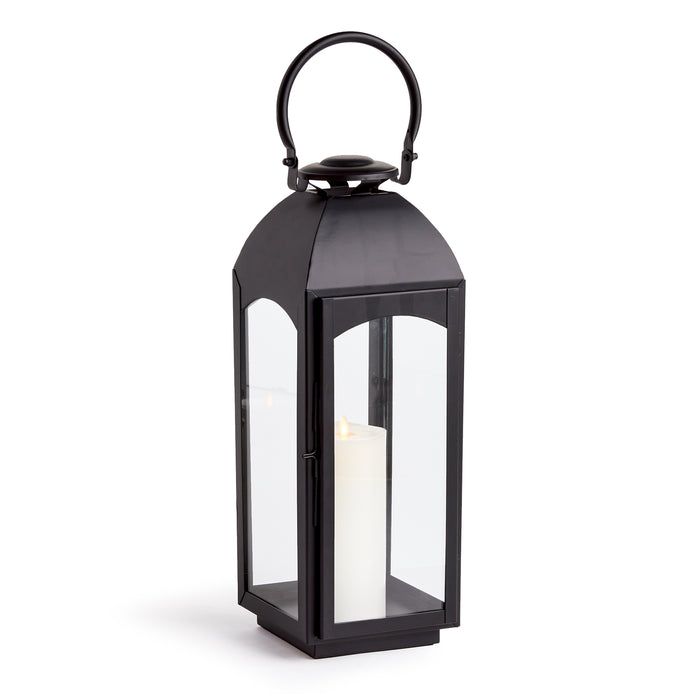 Black metal outdoor lantern with glass panels and a tall white pillar candle inside, arched top handle, weathered finish.