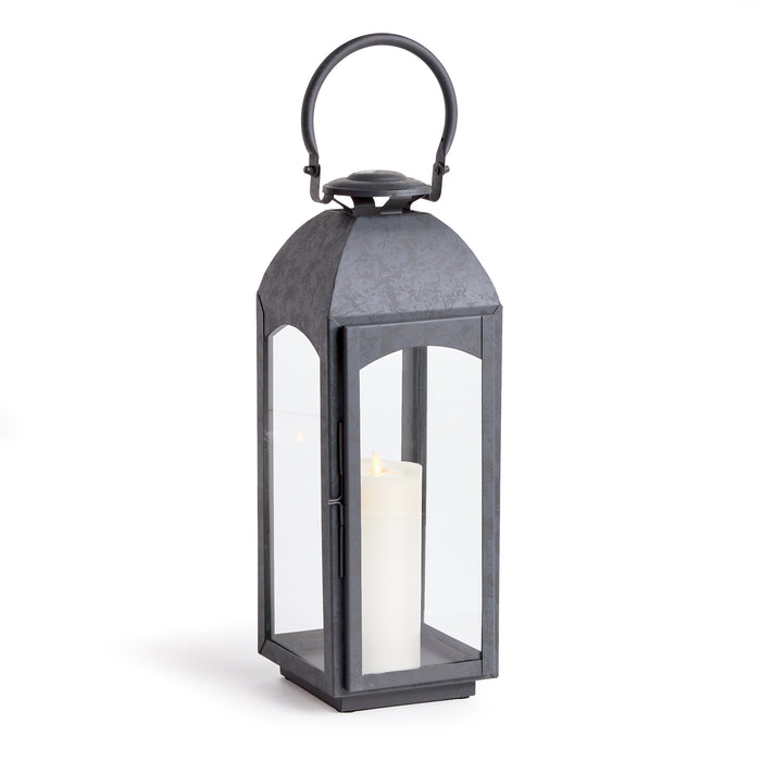 Antoine Outdoor Lantern large, dark metal frame with glass panels and a lit white pillar candle inside, arched handle on top.