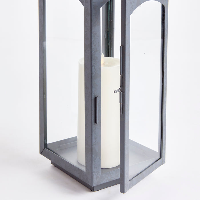 Gray metal outdoor lantern with glass panels enclosing a large white pillar candle inside.