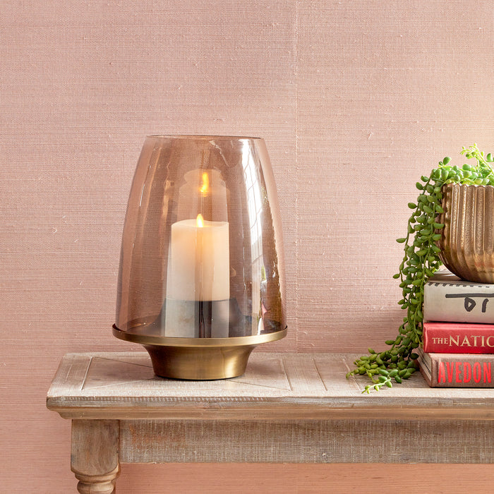 A clear glass hurricane candle holder with a matte brass finish base, displayed on a wooden surface with a bookshelf and potted plant in the background.