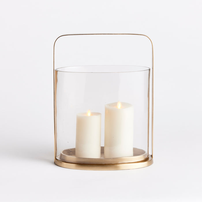 Clear glass hurricane lantern with gold metal handle and base, two white pillar candles inside, neutral backdrop.