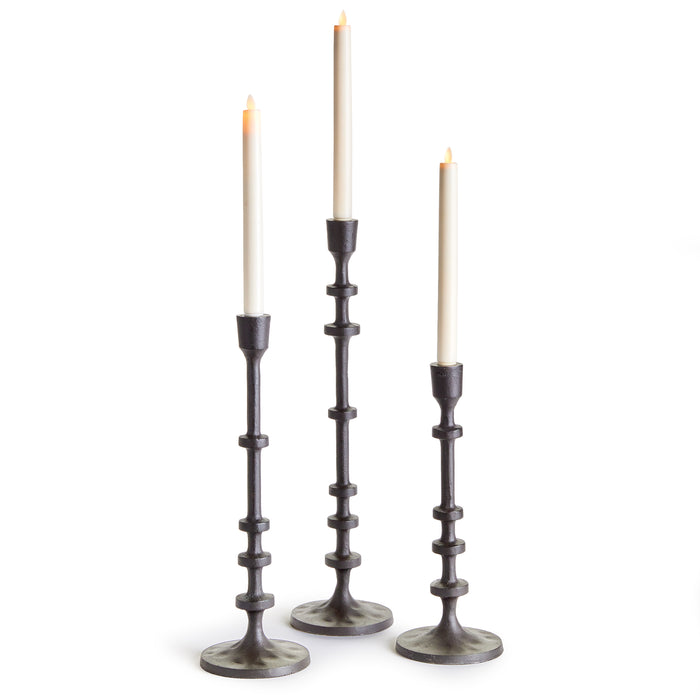 A set of three black tapered candle holders with notched stems, each holding a lit candle.