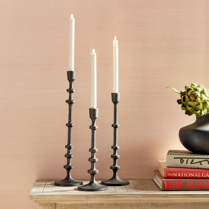 Three dark metal taper holders with white candles on a wooden table, beige backdrop, stack of books, and a green artichoke