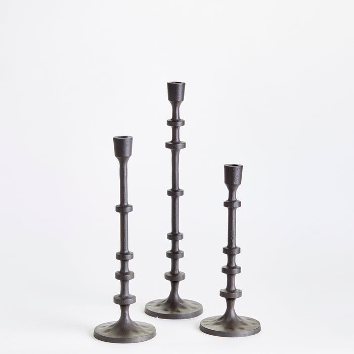 Three dark metal taper holders of varying heights arranged on round bases against a light background.