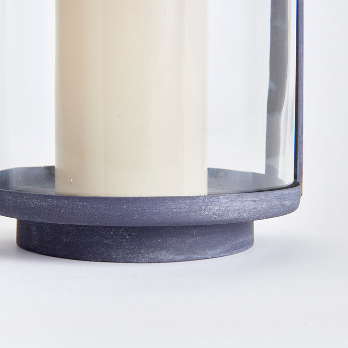 Large outdoor hurricane lantern with a dark blue metal base and cylindrical glass enclosure housing a cream-colored