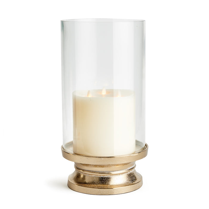 A clear glass footed hurricane candle holder with a snow-on-brass finish, containing a lit candle.