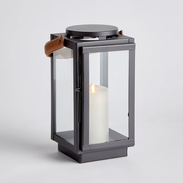 Design Shop Jennings lantern in black metal with glass panels, brown leather strap, and white pillar candle inside, on white