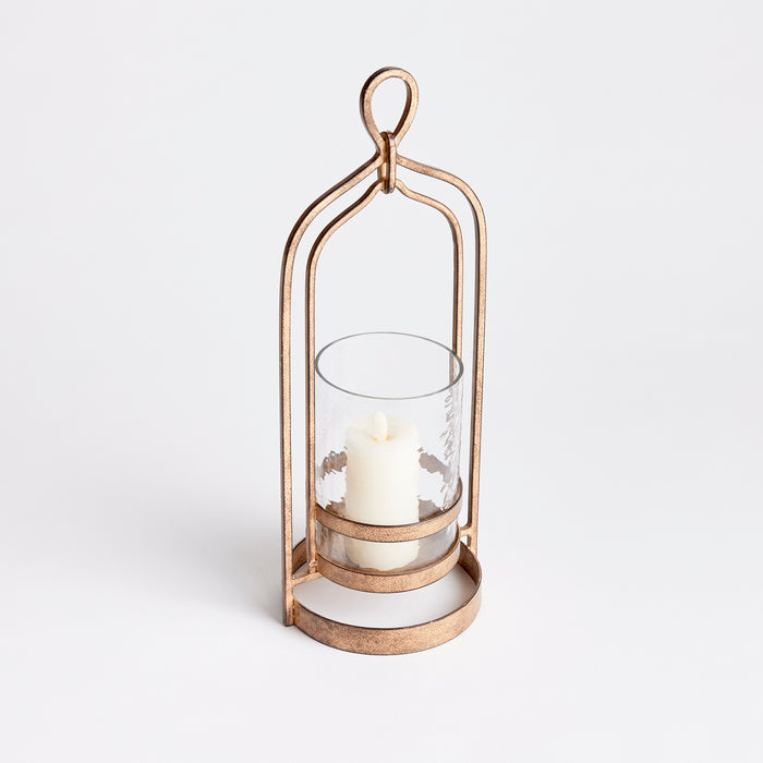 Copper-toned metal hurricane lantern with glass cylinder and white pillar candle, set against a white background.