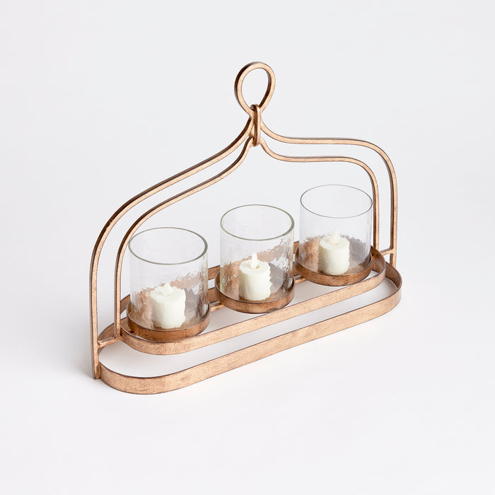 Rose gold metal three-candle hurricane with glass votives on a curved frame and ring handle, empty backdrop.