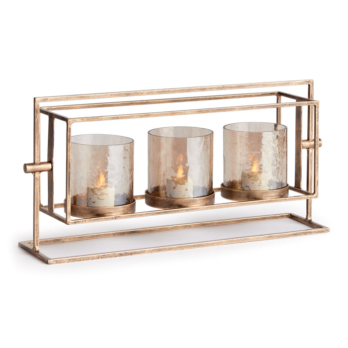 A gold-colored metal candle holder with three glass hurricanes containing candles, featuring a hammered texture and an antique finish.