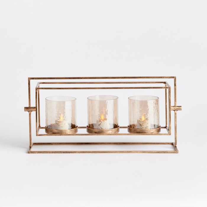 Brass-toned metal three-candle hurricane with cylindrical glass holders and small flickering flames, rectangular frame and