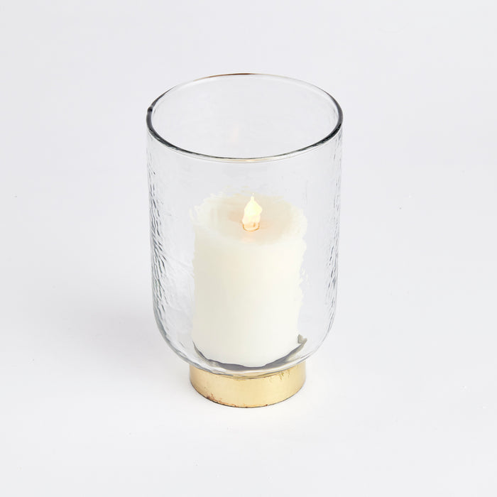 Clear glass hurricane with a white pillar candle, gold-tone base, textured outer wall, plain white background.