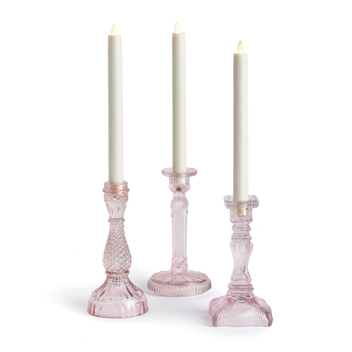 A set of three pink taper candle holders made of glass, each with a unique design, displayed with lit candles inside.