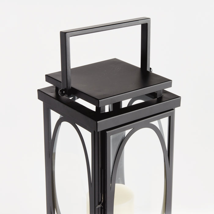 Black metal lantern with glass panels, arched window frames, hinged door, and a rectangular handle on top; candle inside.