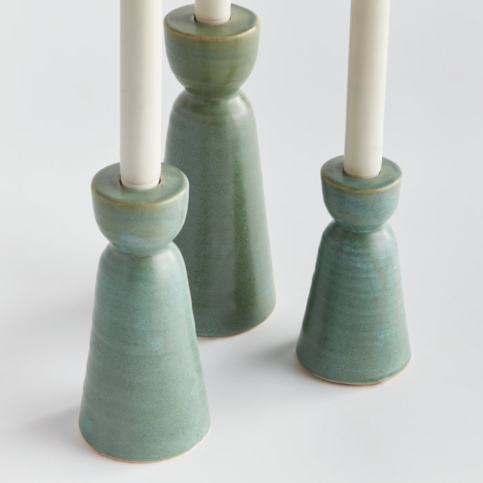 Set of three ceramic green taper candle holders with white candles, earthy glaze, on light background.