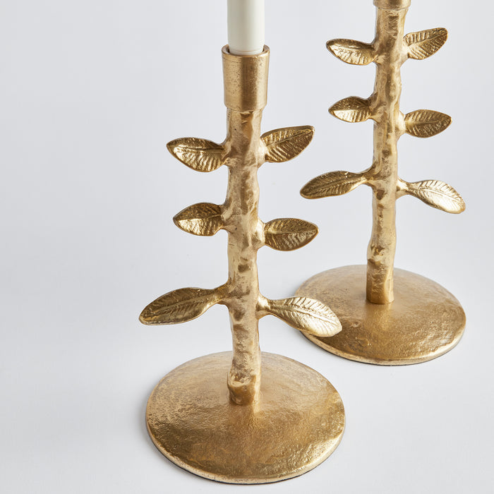 Pair of gold-toned candle holders shaped like branch stems with leaves on circular bases, white candles up top, neutral