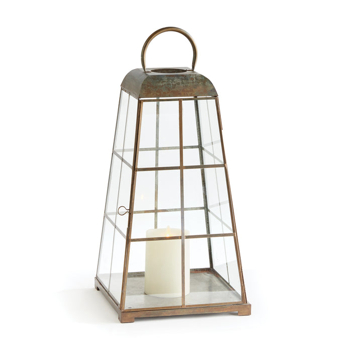 A large lantern with a modern tapered silhouette, featuring upright handles and a warm antique brass finish. It has a glass base and frame, and a candle is lit inside, creating a warm glow.