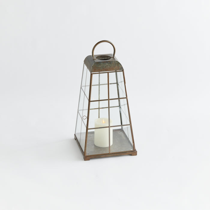 Rusty metal framed lantern with glass panels, arched handle, housing a white pillar candle, set against a white background.