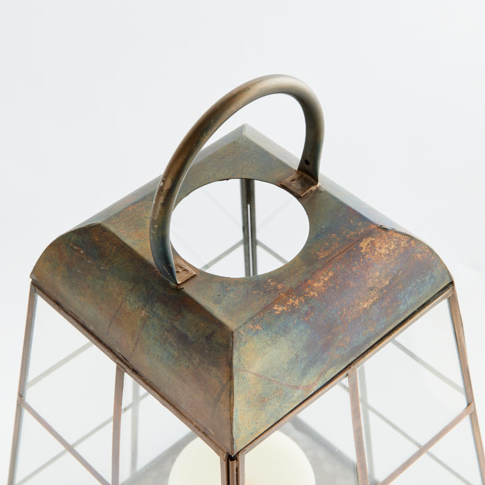 Rusty metal lantern with square frame, curved top handle, open circular chimney slot, weathered patina, candle inside.