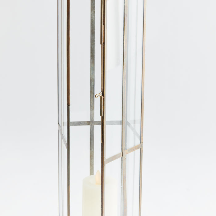 Tall metal-framed lantern with clear glass panels, bronze finish, and a lit pillar candle inside, against a light background.