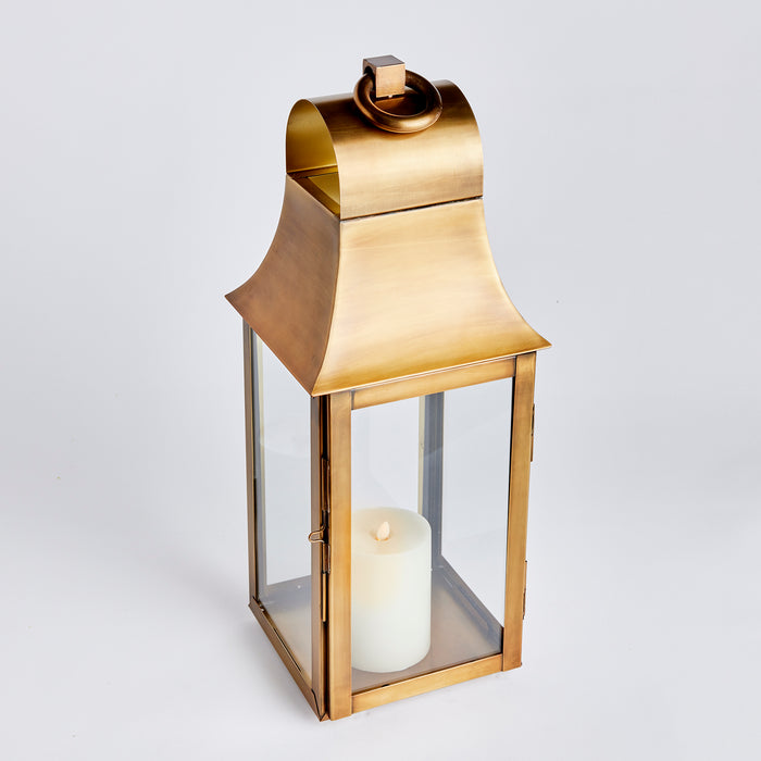Copper-toned metal lantern with glass panels and a white pillar candle inside; small curved top handle and rectangular base