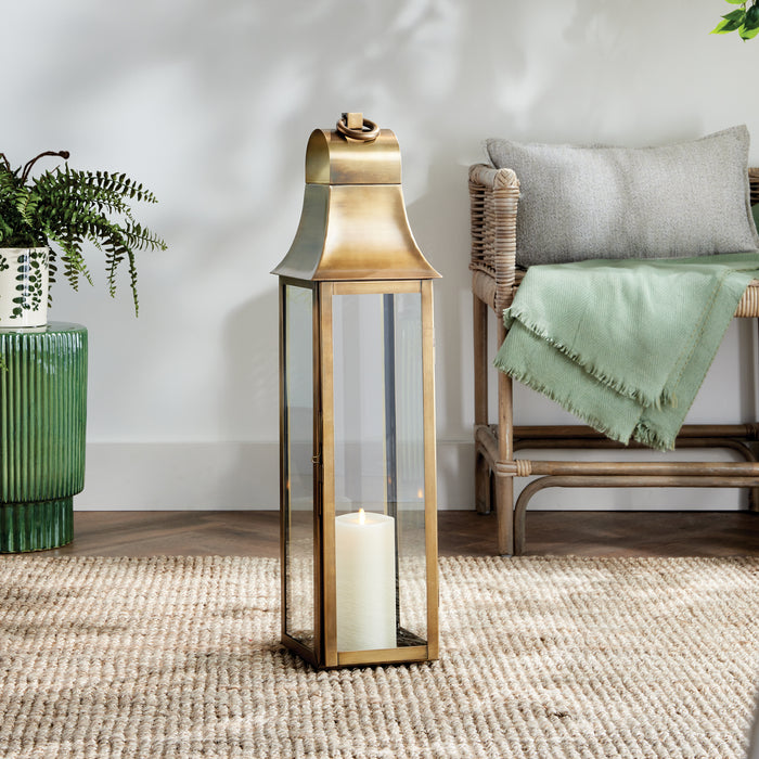 Bronze metal lantern with glass panels and a lit pillar candle inside, set on a woven rug beside a wicker chair.