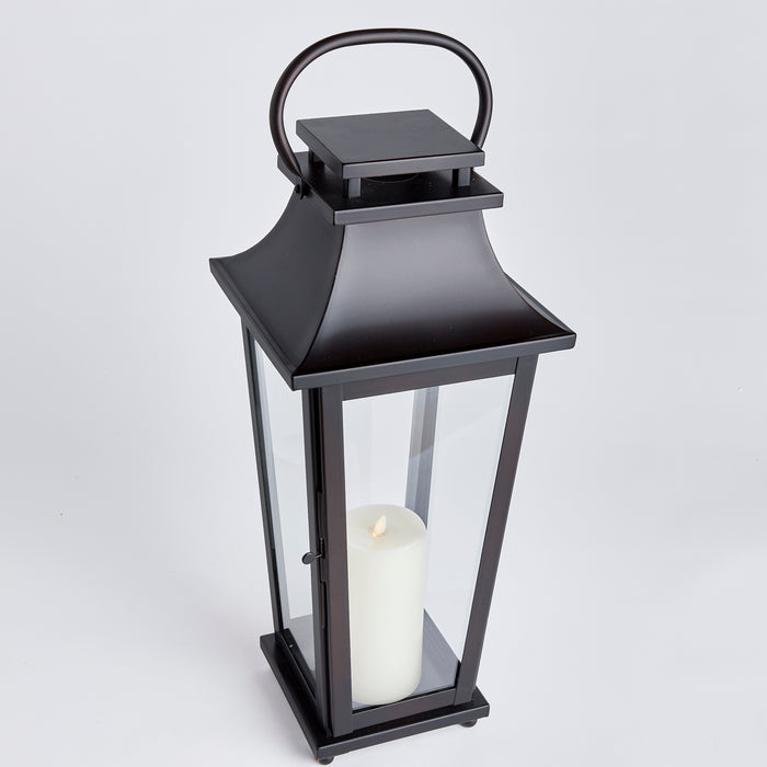 Black outdoor lantern with glass panels, metal frame, curved top roof, and a white pillar candle inside.