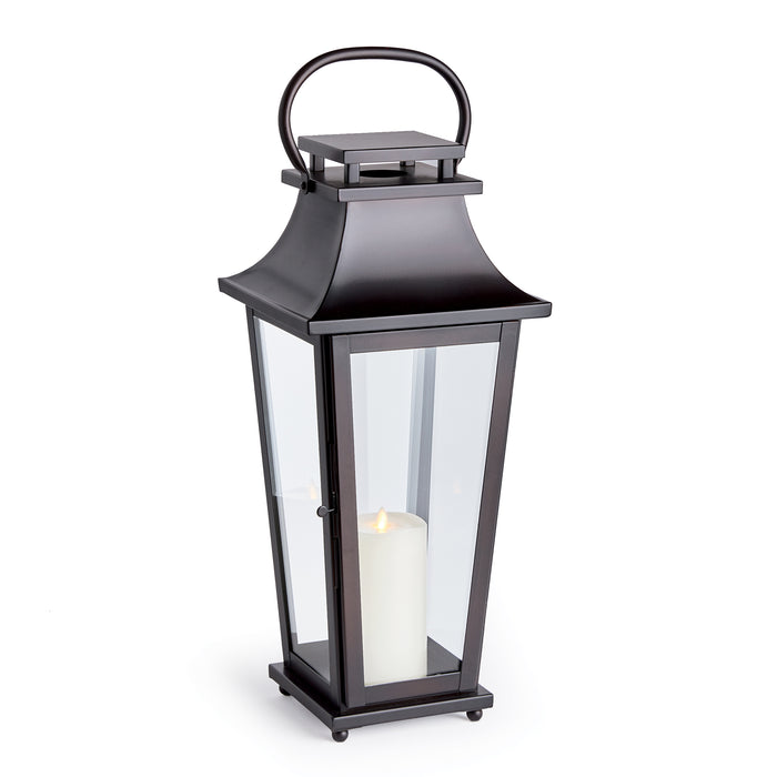 Black outdoor lantern with glass panels, metal frame, curved top loop, and white pillar candle inside on a square base.