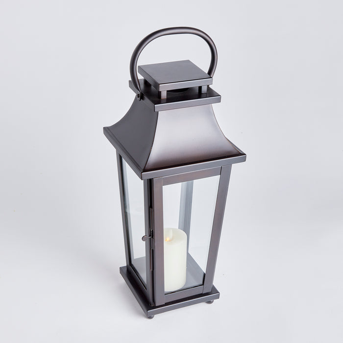 Outdoor metal lantern with glass panels, dark gray finish, curved top handle, pillar candle inside, set against a light