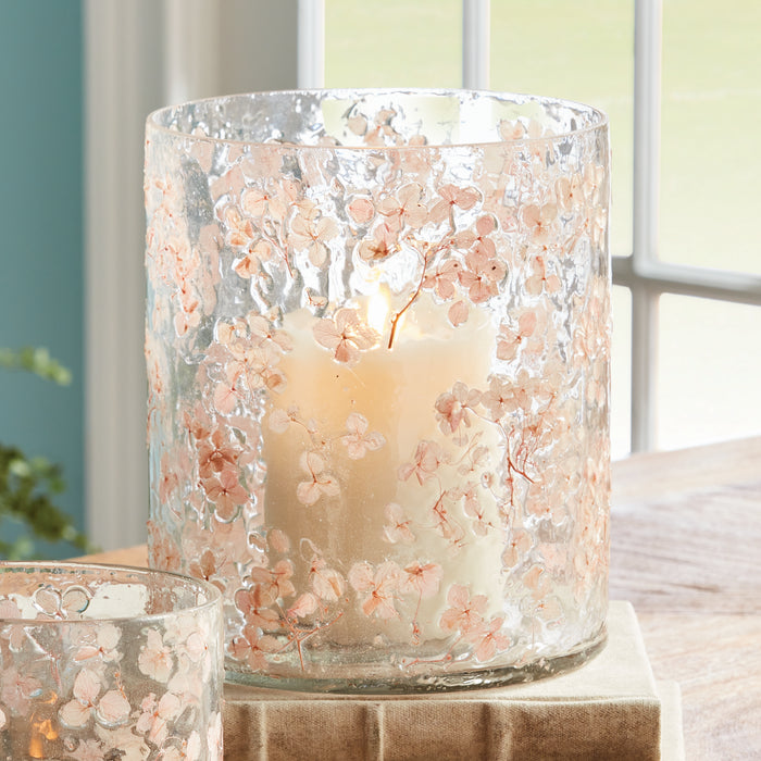 A frosted glass hurricane with pink cherry blossom floral pattern and a lit pillar candle inside on a beige fabric base by a